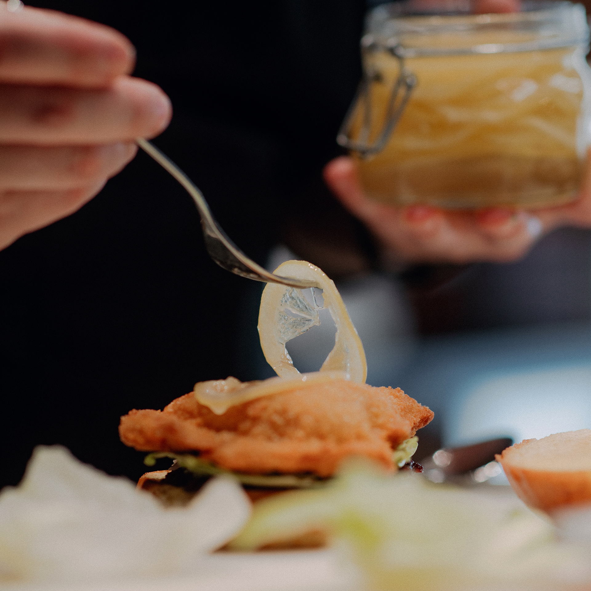 Mit einer Gabel wird ein Stück eingelegte Zitrone auf ein paniertes Schnitzel im Burger gelegt; im Hintergrund ein Glas mit Zitronen.