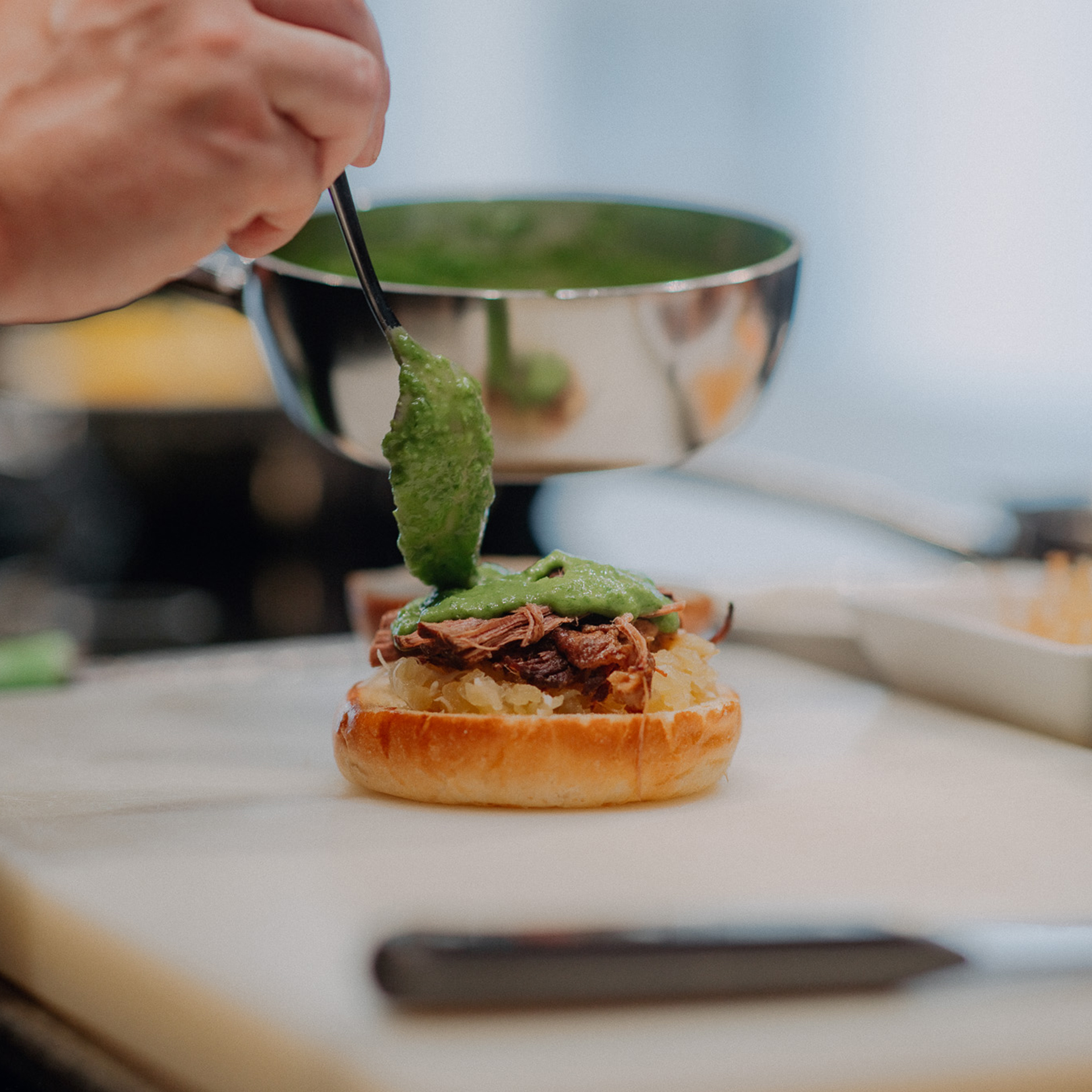 Eine Hand löffelt grüne Schnittlauchsauce aus einer Metallschüssel auf einen Burger mit Fleisch und Kraut.