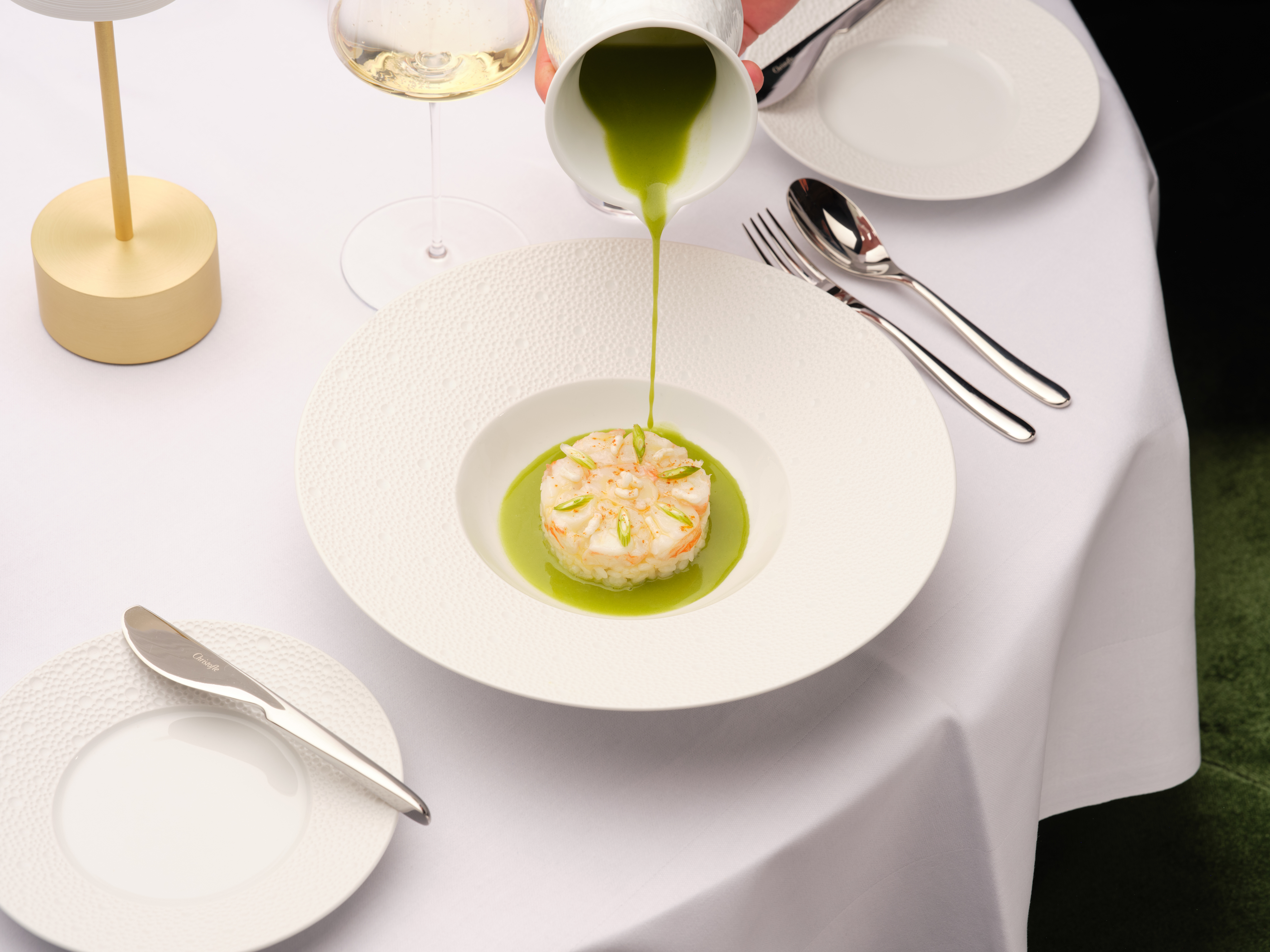Aus einem kleinen Krug wird leuchtend grüne Sauce über eine fein angerichtete Vorspeise in einem tiefen weißen Teller gegossen.