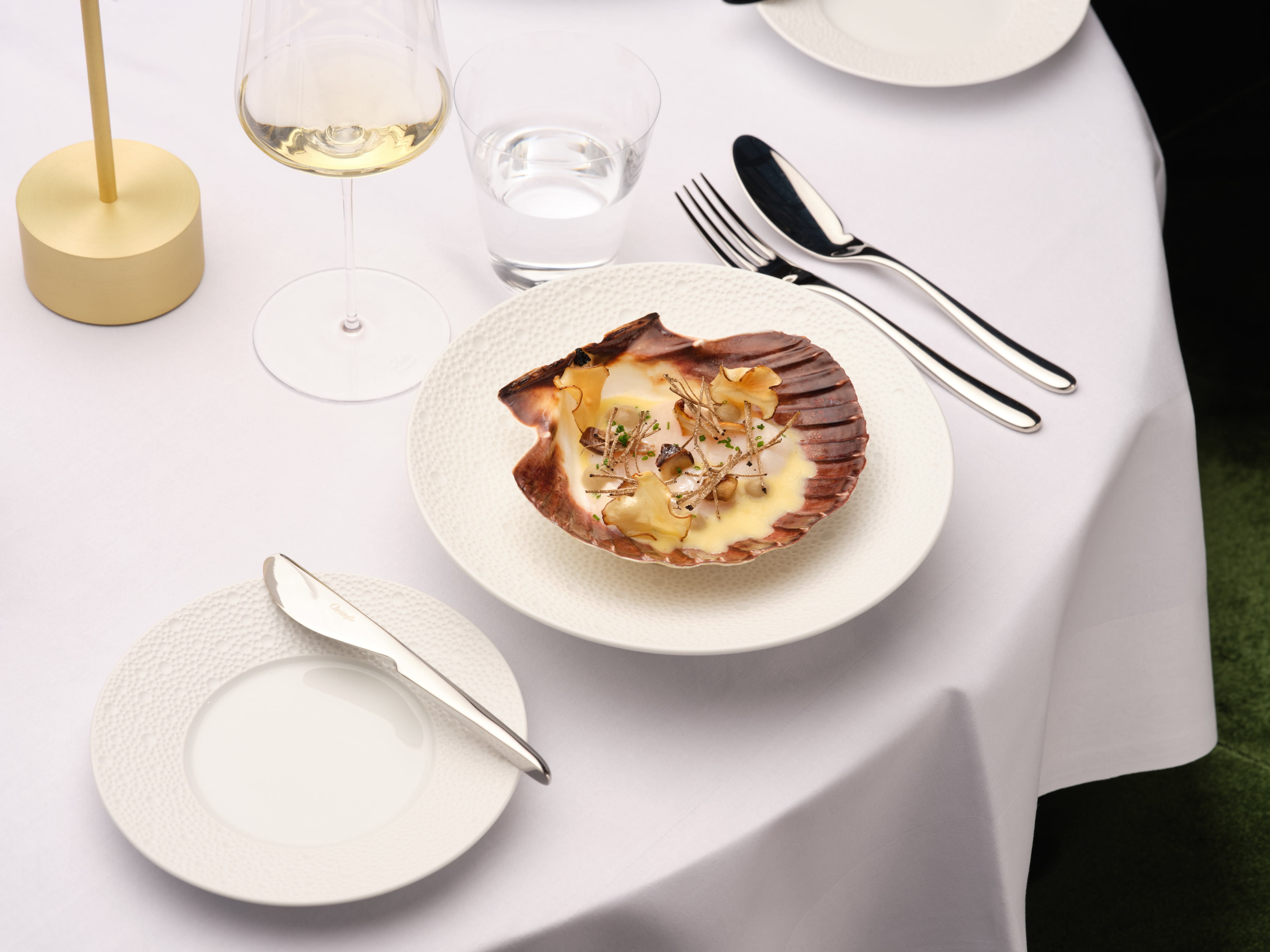 Eine kunstvoll angerichtete Jakobsmuschel liegt in ihrer Schale auf einem weißen Teller, gedeckt auf einer eleganten Tafel mit Wein- und Wasserglas.