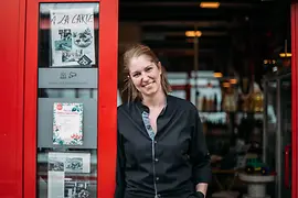 A woman in black workwear stands in the entrance of a venue with a red door frame, looking at the camera.