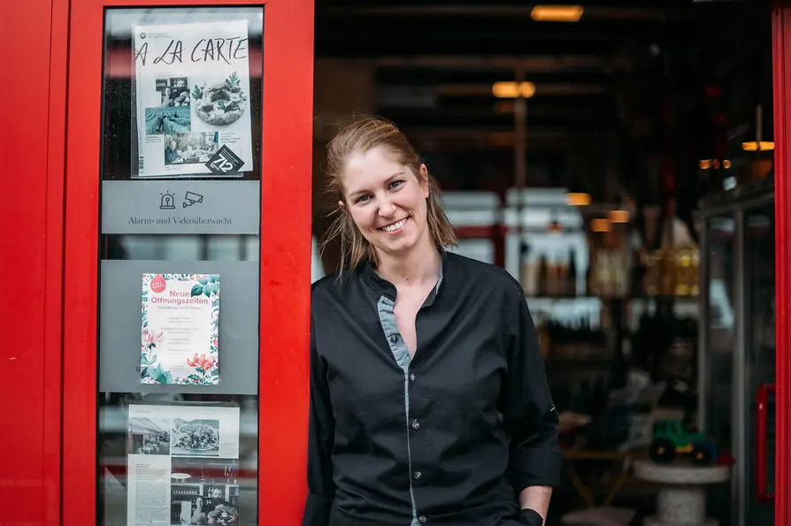A woman in black workwear stands in the entrance of a venue with a red door frame, looking at the camera.