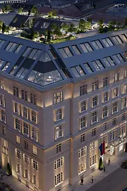 A large, elegantly illuminated hotel building in Vienna seen from above at dusk. The rooftop features several landscaped terraces with trees, seating areas, and warm lighting. Surrounding streets and historic neighboring buildings are visible.