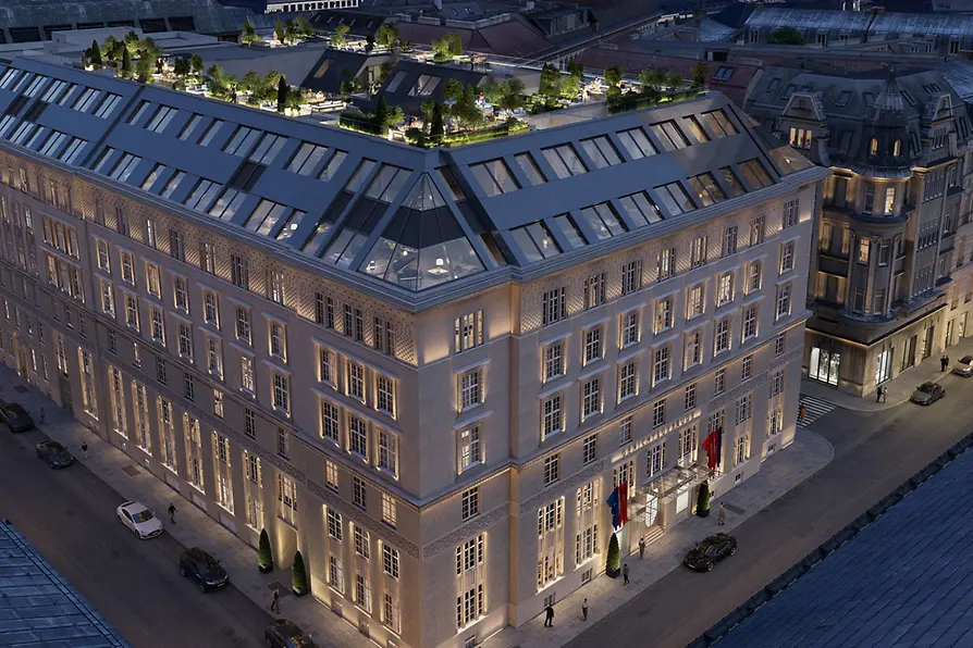 A large, elegantly illuminated hotel building in Vienna seen from above at dusk. The rooftop features several landscaped terraces with trees, seating areas, and warm lighting. Surrounding streets and historic neighboring buildings are visible.