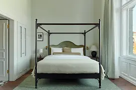 A serene suite bedroom featuring a four-poster bed with soft green upholstery, tall windows with flowing curtains, and minimalist bedside furnishings.
