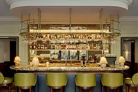 An elegant hotel bar with a curved marble counter, olive-green bar stools, brass shelving, and a backlit display of bottles and glassware.