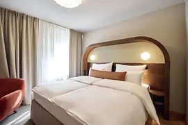 A bright hotel room featuring a neatly made double bed with a wooden arched headboard, warm lighting, long beige curtains, and an orange upholstered armchair.