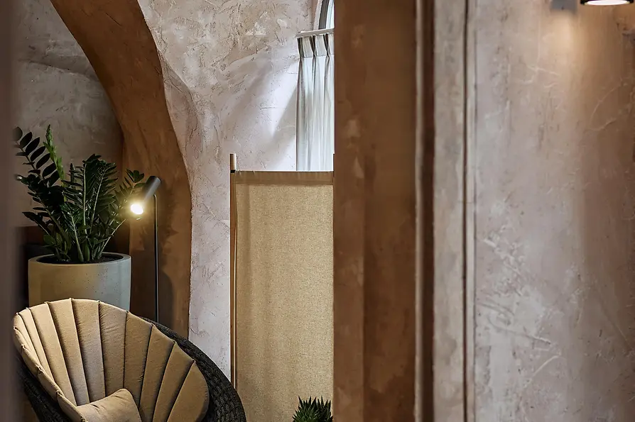 Ein sonniger Innenhof mit modernen Outdoor-Sitzmöbeln, gepolsterten Sesseln um einen kleinen Tisch und einem gepflegten Baum, der Schatten auf die gelbe Fassade mit Rundbogenfenstern wirft.