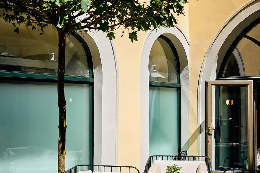 Eine gemütliche Lounge-Ecke mit einem gepolsterten, geflochtenen Schaukelstuhl, kleinen runden Tischen und Topfpflanzen vor strukturierten Steinwänden und sanfter Beleuchtung.