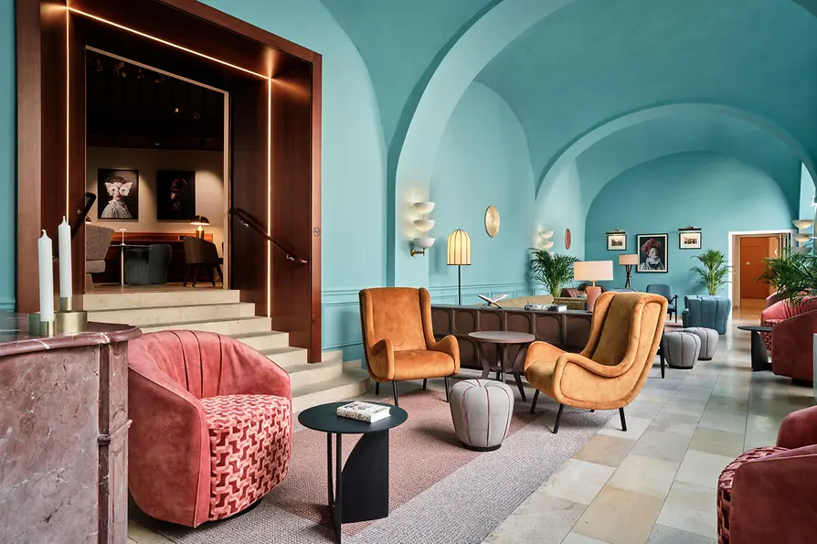 Eine farbenfrohe Lounge mit türkisfarbenen Gewölbedecken, farbenfrohen Sitzmöbeln im Mid-Century-Stil, sanfter Beleuchtung und gerahmter Kunst – lebhaft und zugleich elegant.