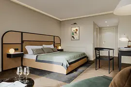 A bright and elegant hotel room featuring a large bed with a cane-and-leather headboard, minimalistic black furniture, soft neutral walls, and a framed artwork above the bed.