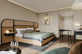 A bright and elegant hotel room featuring a large bed with a cane-and-leather headboard, minimalistic black furniture, soft neutral walls, and a framed artwork above the bed.