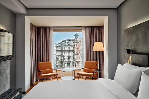 A modern hotel room with a large bed facing a wide picture window, two retro-style orange armchairs, and a view of historic Vienna buildings outside.
