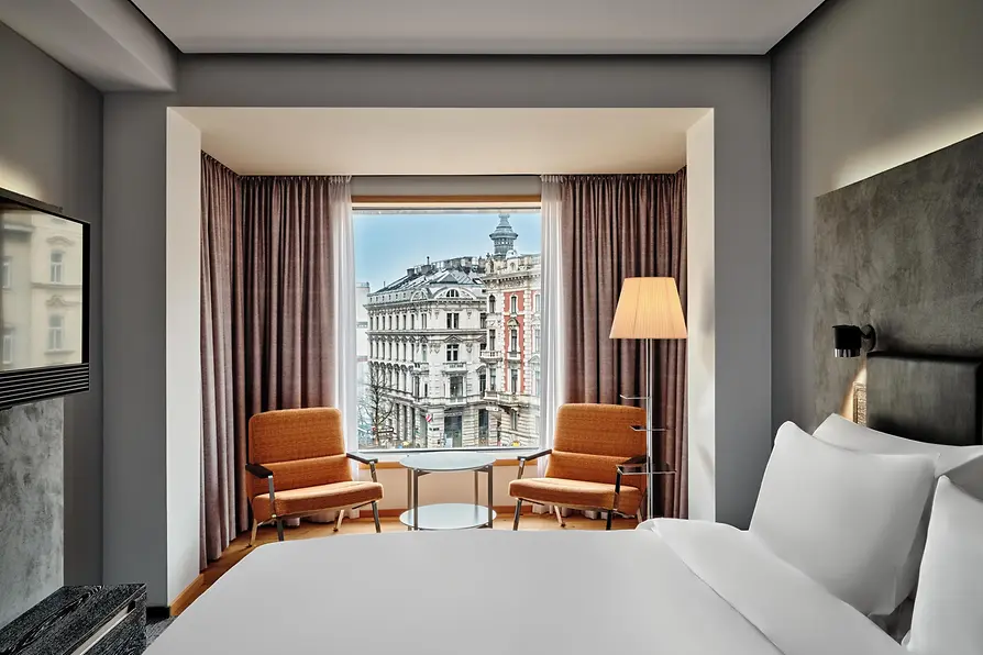 A modern hotel room with a large bed facing a wide picture window, two retro-style orange armchairs, and a view of historic Vienna buildings outside.
