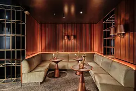 A stylish, dimly lit bistro lounge with warm wooden wall panels, soft corner seating, copper-toned tables, and ambient lighting creating an intimate atmosphere.