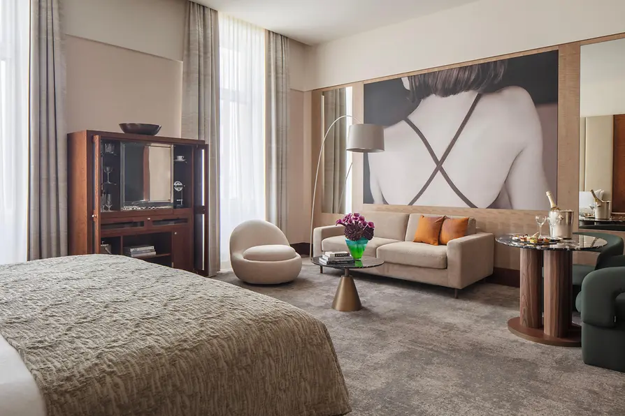 A spacious, elegant hotel suite with a large bed, modern seating, marble tables, and a striking wall-sized artwork of a woman’s back, complemented by natural light from tall windows.