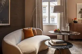 A stylish hotel lounge area featuring a curved cream sofa with orange cushions, marble coffee tables, and a window letting in soft daylight, with fruit and glassware arranged neatly on the table.