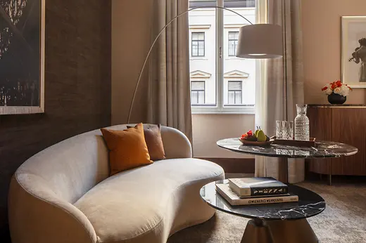 A stylish hotel lounge area featuring a curved cream sofa with orange cushions, marble coffee tables, and a window letting in soft daylight, with fruit and glassware arranged neatly on the table.