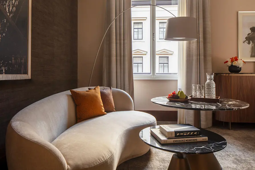 Ein stilvoller Lounge-Bereich eines Hotelzimmers mit geschwungenem cremefarbenem Sofa und orangefarbenen Kissen, Marmorcouchtischen und einem Fenster, das sanftes Tageslicht hereinlässt; auf dem Tisch stehen Obst und Glaswaren.