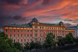 The historic façade of the Anantara Palais Hansen Vienna glows warmly at sunset, with a dramatic sky of red and purple clouds above the grand building lined with trees.