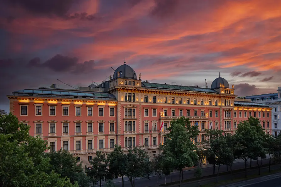 Die historische Fassade des Anantara Palais Hansen Vienna leuchtet im warmen Licht des Sonnenuntergangs, während ein dramatischer Himmel mit roten und violetten Wolken über dem prachtvollen, von Bäumen gesäumten Gebäude steht.