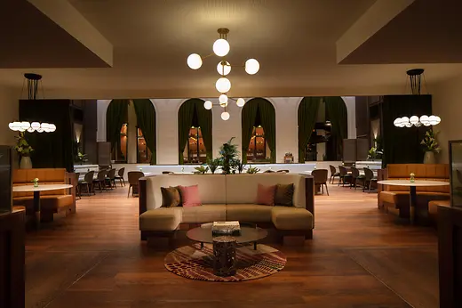 A warmly lit brasserie with elegant mid-century–style seating, green curtains, round hanging lamps, and a central curved sofa arranged around a low marble table.
