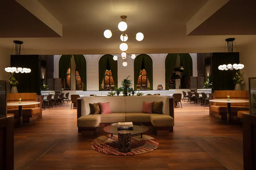 A warmly lit brasserie with elegant mid-century–style seating, green curtains, round hanging lamps, and a central curved sofa arranged around a low marble table.