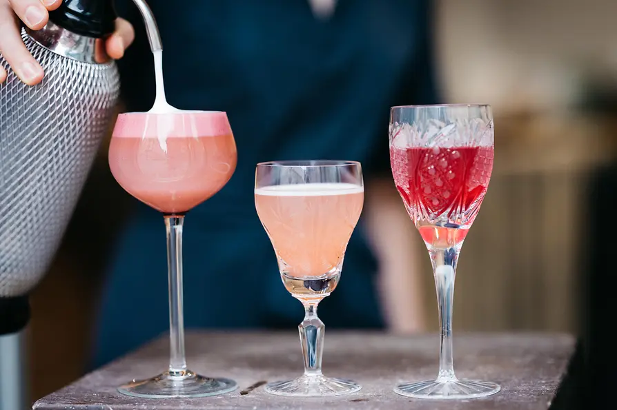 Eine Person gießt Schaum auf einen rosafarbenen Cocktail. Drei farbenfrohe Drinks in verschiedenen Gläsern stehen nebeneinander, von schaumigem Rosa bis zu tiefem Rot.