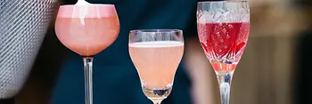 Eine Person gießt Schaum auf einen rosafarbenen Cocktail. Drei farbenfrohe Drinks in verschiedenen Gläsern stehen nebeneinander, von schaumigem Rosa bis zu tiefem Rot.