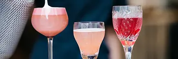 Eine Person gießt Schaum auf einen rosafarbenen Cocktail. Drei farbenfrohe Drinks in verschiedenen Gläsern stehen nebeneinander, von schaumigem Rosa bis zu tiefem Rot.