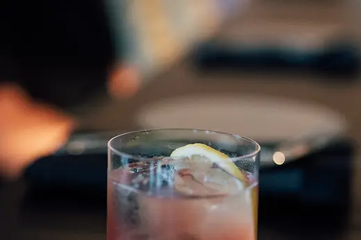 A close-up of a pink spritz cocktail with ice cubes and a lemon slice in a clear glass, set against a softly blurred bar background.