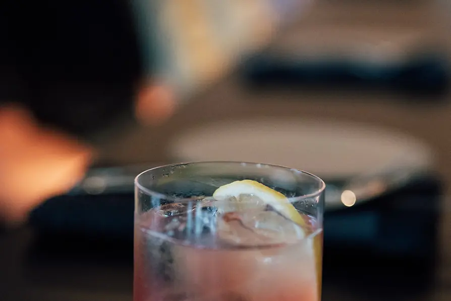 A close-up of a pink spritz cocktail with ice cubes and a lemon slice in a clear glass, set against a softly blurred bar background.