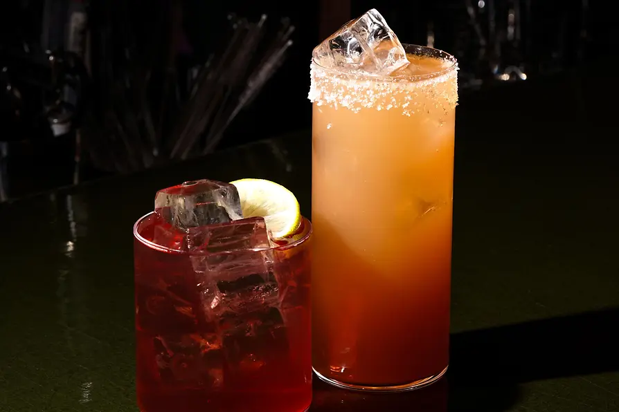Two cocktails on a bar counter: a short red drink with large ice cubes and a lemon slice, and a tall orange cocktail with a salted rim and ice.