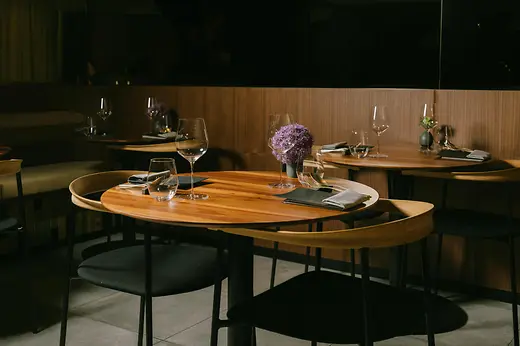 A warmly lit restaurant interior with wooden tables, elegant glassware, black chairs, and purple floral centerpieces.