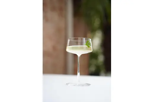 A tall, elegant coupe glass filled with a pale green cocktail, garnished with a fresh parsley leaf, set on a white tablecloth with soft greenery blurred in the background.