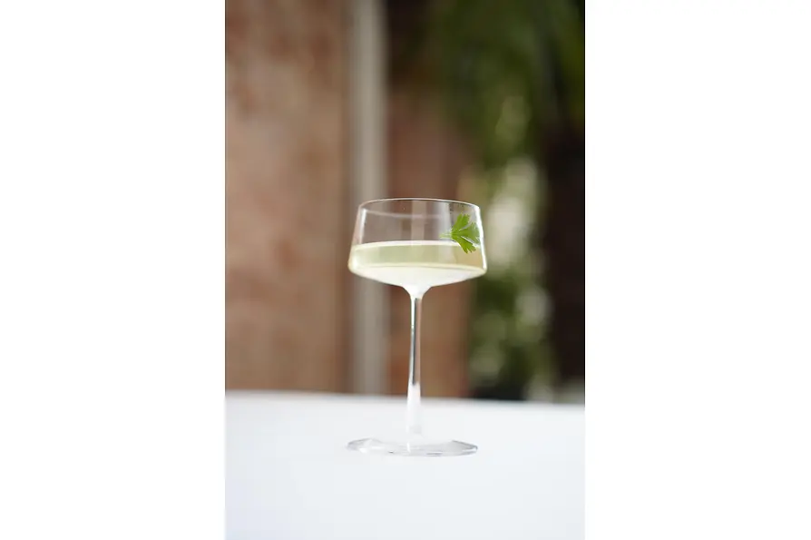Ein hohes, elegantes Coupé-Glas mit einem hellgrünen Cocktail, garniert mit einem frischen Petersilienblatt, auf einem weißen Tischtuch mit weich verschwommenem Grün im Hintergrund.