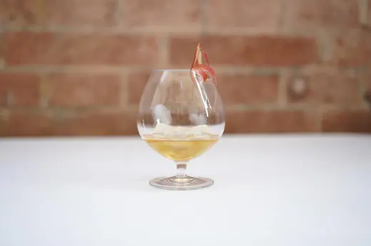 A rounded glass with a golden cocktail on ice, garnished with a thin dried apple slice, placed on a white tablecloth with a blurred brick wall behind.
