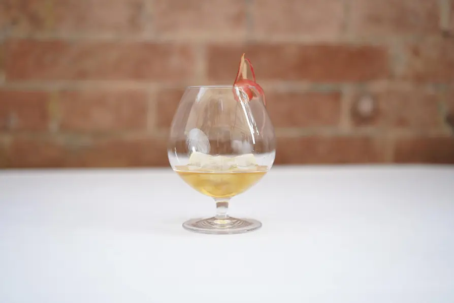 A rounded glass with a golden cocktail on ice, garnished with a thin dried apple slice, placed on a white tablecloth with a blurred brick wall behind.