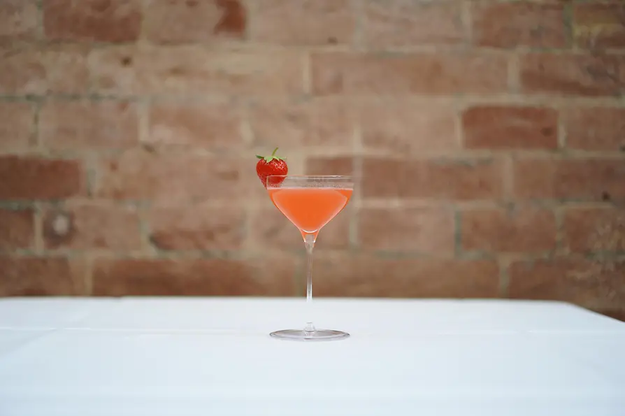 Ein rosafarbener Cocktail in einem hohen Stielglas, garniert mit einer frischen Erdbeere am Glasrand, auf einem weißen Tischtuch vor einer unscharfen Backsteinwand.