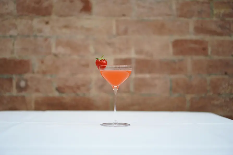 A pink cocktail served in a tall stemmed glass, garnished with a fresh strawberry on the rim, placed on a white tablecloth with a blurred brick wall in the background.