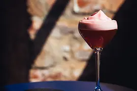 Ein hochstieliges Glas mit einem dunkelroten Cocktail und einer dicken rosafarbenen Schaumschicht, dramatisch beleuchtet vor einer strukturierten Ziegelwand.