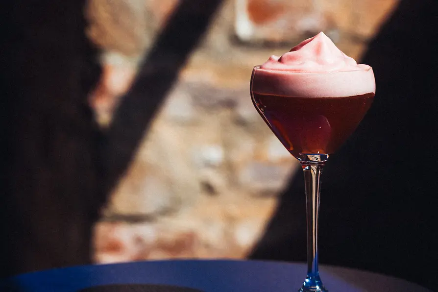 A tall-stemmed glass with a dark red cocktail topped with a thick pink foam, illuminated dramatically against a textured brick wall.