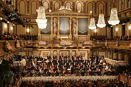 A grand view of the Vienna Musikverein’s Golden Hall during the New Year’s Concert, with the orchestra performing on a flower-decorated stage, surrounded by golden balconies filled with an audience and large crystal chandeliers.