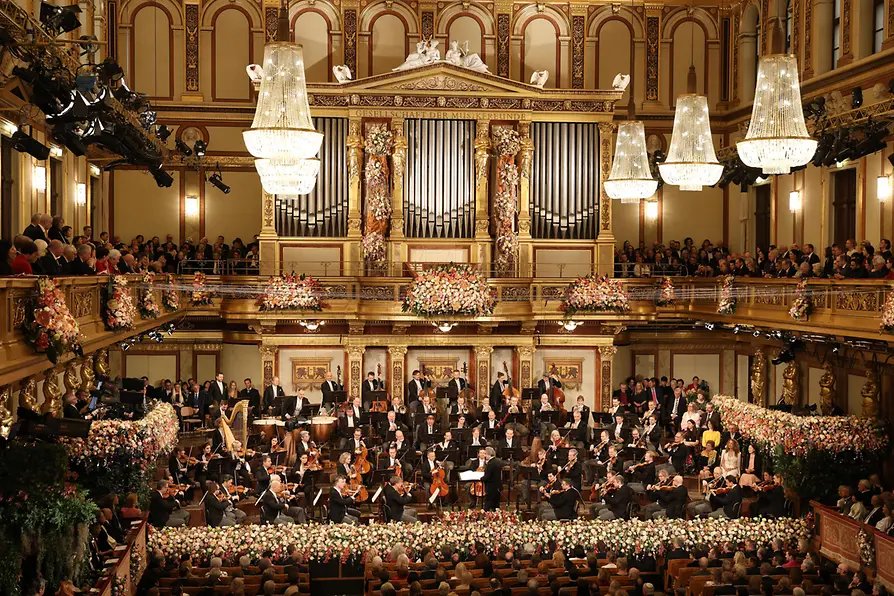 A grand view of the Vienna Musikverein’s Golden Hall during the New Year’s Concert, with the orchestra performing on a flower-decorated stage, surrounded by golden balconies filled with an audience and large crystal chandeliers.