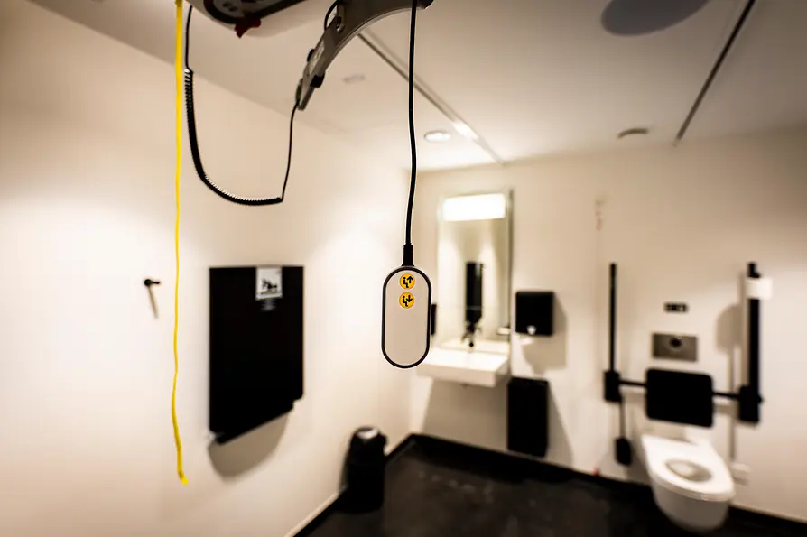 Innenraum einer barrierefreien Toilette mit deckenmontiertem Hebesystem und hängender Fernbedienung, einem Waschbecken mit Spiegel sowie Stützgriffen neben dem WC.