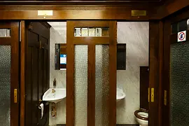 Innenansicht einer historischen öffentlichen Toilette mit dunklen Holztüren, Milchglasfenstern, gemustertem Fliesenboden und Vintage-Waschbecken vor Marmortafeln.