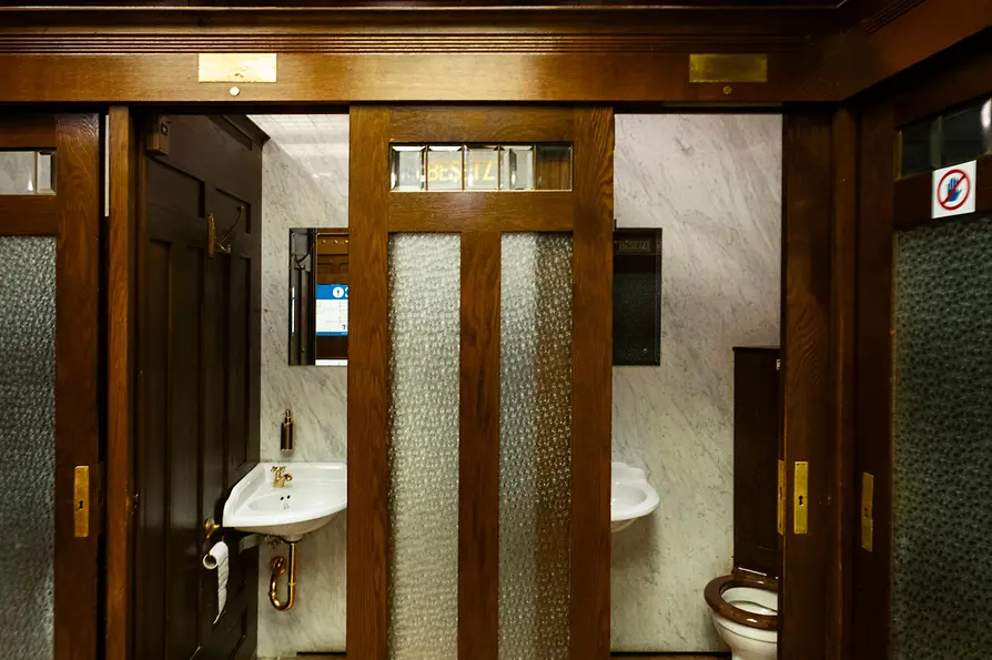 Innenansicht einer historischen öffentlichen Toilette mit dunklen Holztüren, Milchglasfenstern, gemustertem Fliesenboden und Vintage-Waschbecken vor Marmortafeln.