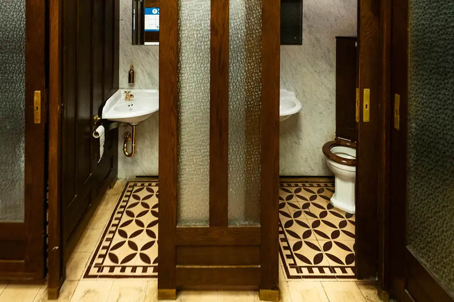 Innenansicht einer historischen öffentlichen Toilette mit dunklen Holztüren, Milchglasfenstern, gemustertem Fliesenboden und Vintage-Waschbecken vor Marmortafeln.
