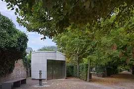 Ein modernes öffentliches WC-Gebäude mit metallischer Fassade entlang eines baumbestandenen Weges im Augarten. Laub liegt teilweise am Boden, Bänke und Hecken rahmen die ruhige Parksituation ein.