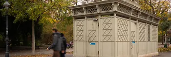 A historic light-gray public restroom pavilion with decorative lattice panels and stained-glass details, set among autumn trees as pedestrians walk by.